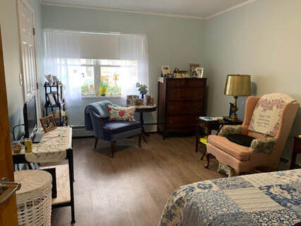 Photo of a bedroom. In the foreground is a bed with a blue and white patchwork bedspread on it. Across from the bed is a table with small television and family photographs. Toward the back of the room, in front of the large double window, is a set of upholstered chairs, two side tables, a corner curio and a tall bureau.